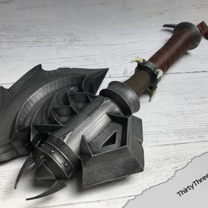 May include: A large, detailed, silver-colored axe with a leather-wrapped handle. The axe head has a complex design with sharp points and a textured surface. The handle has a brown leather wrap and a metal ring at the top.