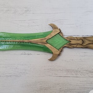 May include: A green and gold decorative sword with a detailed design. The blade is green and the hilt is gold with intricate details.
