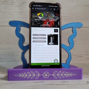 Peut inclure: Un support de t&eacute;l&eacute;phone en bois violet avec un motif de papillon bleu. Le support a un motif floral sur le devant. Un smartphone est dans le support affichant le site Web "thirtythree3d.com". Le site Web a une barre de recherche, des boutons pour "Boutique", "Nous contacter" et "Photos des clients". Le site Web a &eacute;galement une section intitul&eacute;e "Impression 3D".