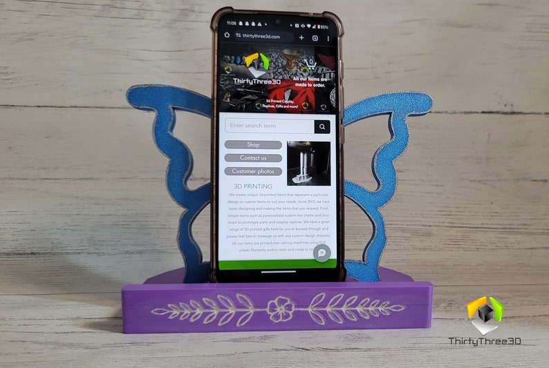 Peut inclure: Un support de t&eacute;l&eacute;phone en bois violet avec un motif de papillon bleu. Le support a un motif floral sur le devant. Un smartphone est dans le support affichant le site Web "thirtythree3d.com". Le site Web a une barre de recherche, des boutons pour "Boutique", "Nous contacter" et "Photos des clients". Le site Web a &eacute;galement une section intitul&eacute;e "Impression 3D".