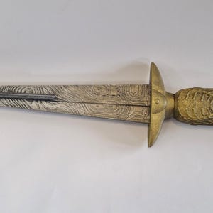 May include: A decorative dagger with a patterned blade and a gold-colored handle. The handle features an ornate design and a red gem at the pommel. The blade has a textured, wood-grain appearance.