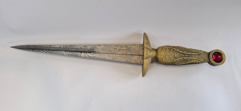 May include: A decorative dagger with a patterned blade and a gold-colored handle. The handle features an ornate design and a red gem at the pommel. The blade has a textured, wood-grain appearance.