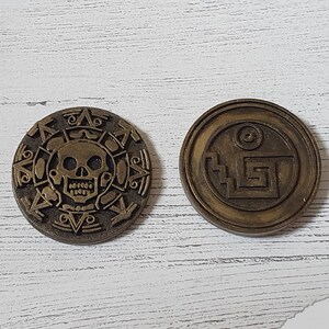 May include: Two gold-colored metal coins with intricate designs. One coin features a skull and crossbones, while the other has a geometric pattern.
