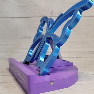 Peut inclure: Un support de t&eacute;l&eacute;phone violet avec un motif de papillon bleu. Le support a un petit bouton sur le devant et une rainure pour tenir un t&eacute;l&eacute;phone.