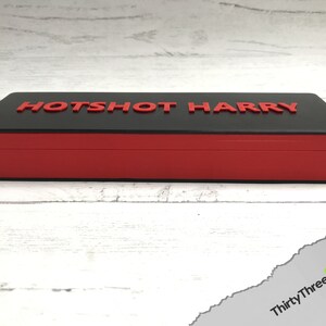 Pode incluir: Uma caixa retangular preta e vermelha com as palavras "HOTSHOT HARRY" impressas em vermelho na parte superior.
