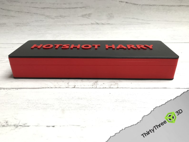 Pode incluir: Uma caixa retangular preta e vermelha com as palavras "HOTSHOT HARRY" impressas em vermelho na parte superior.