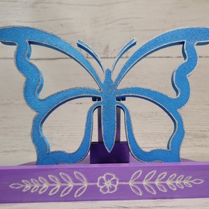 Peut inclure: Une figurine de papillon bleu avec des accents de paillettes argent&eacute;es, pos&eacute;e sur un socle violet avec un motif floral blanc. Le socle est orn&eacute; d'un motif floral blanc.