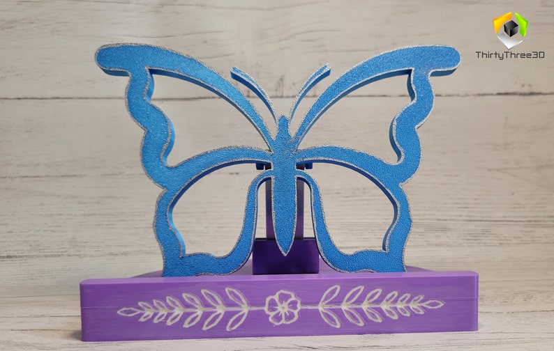 Peut inclure: Une figurine de papillon bleu avec des accents de paillettes argent&eacute;es, pos&eacute;e sur un socle violet avec un motif floral blanc. Le socle est orn&eacute; d'un motif floral blanc.