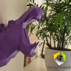 May include: Purple 3D printed geometric hippopotamus head wall decor. The hippopotamus is facing the right side of the image with its mouth open. The hippopotamus is mounted on a white wall.