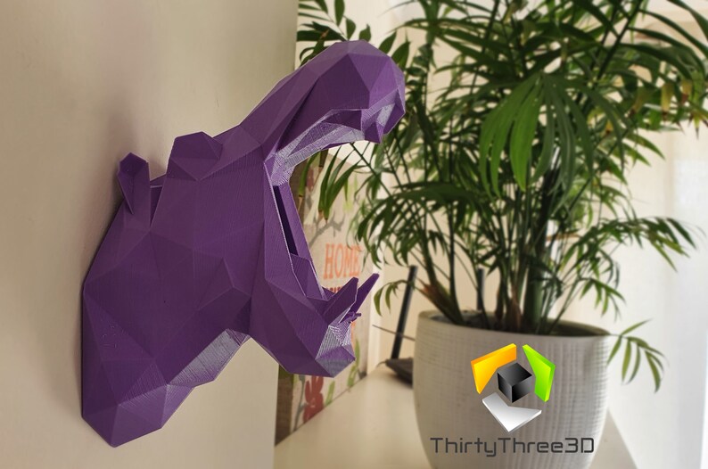May include: Purple 3D printed geometric hippopotamus head wall decor. The hippopotamus is facing the right side of the image with its mouth open. The hippopotamus is mounted on a white wall.