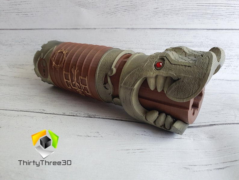 May include: A 3D printed lightsaber hilt with a dragon head design. The hilt is brown and has a red gem for an eye. The dragon's mouth is open and has sharp teeth.