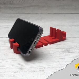 Op de afbeelding: Een rode 3D-geprinte telefoonstandaard in de vorm van het woord "TIME". Een zwarte smartphone rust op de standaard.