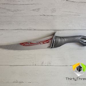 May include: A silver and red dagger with a black handle and a red design on the blade. The dagger is on a white wooden surface.