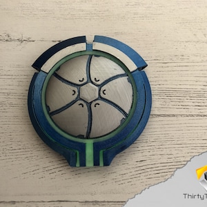 May include: A 3D printed, blue, green, and white circular object with a geometric design. The object has a raised center with a hexagonal pattern.