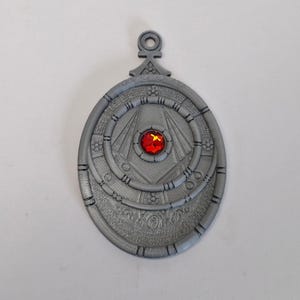 May include: A silver-colored pendant with an oval shape and intricate engraved details. The pendant features a central red gemstone and a loop at the top for hanging. The design includes concentric circles and geometric patterns.