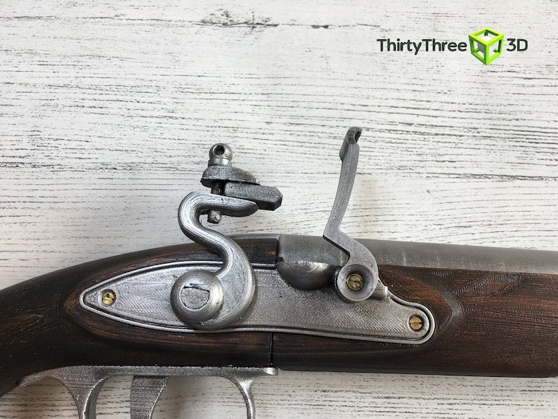 Puede incluir: Primer plano de una r&eacute;plica de pistola de chispa. La pistola presenta una culata de madera marr&oacute;n oscura, una cerradura de metal plateado y un mecanismo de gatillo. El fondo es de madera blanca.