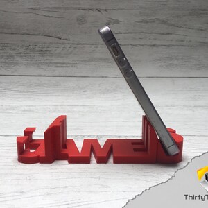 May include: A red 3D printed phone stand shaped like the word "GAMES". A silver smartphone is propped up in the stand.