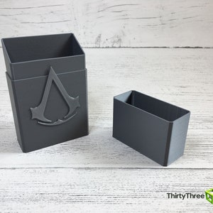 May include: A gray plastic box with a raised Assassin's Creed logo on the front. The box is open and a smaller gray plastic box is sitting next to it.