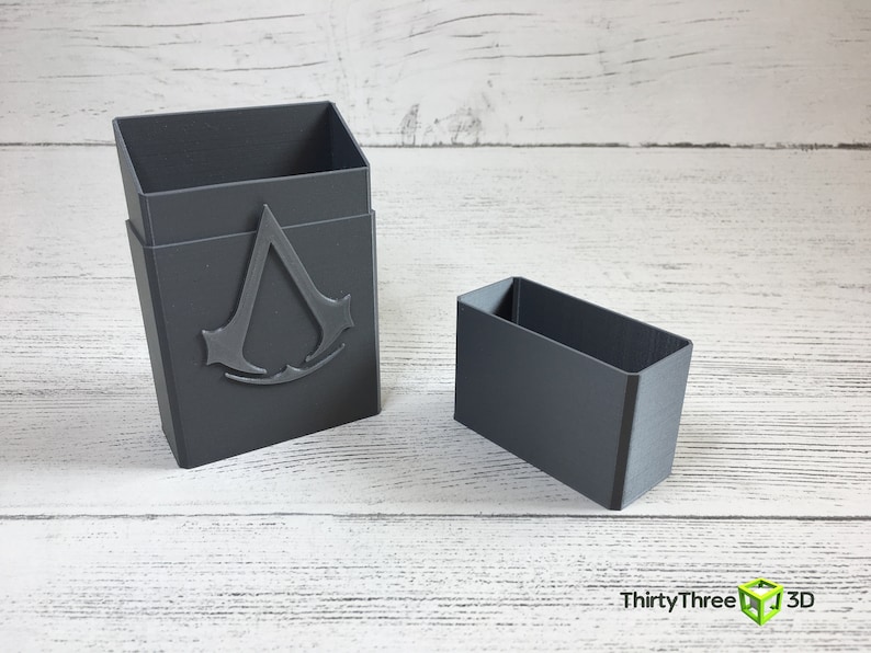May include: A gray plastic box with a raised Assassin's Creed logo on the front. The box is open and a smaller gray plastic box is sitting next to it.