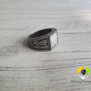 May include: A silver ring with a square face and intricate designs. The ring has a beaded edge and a textured surface.