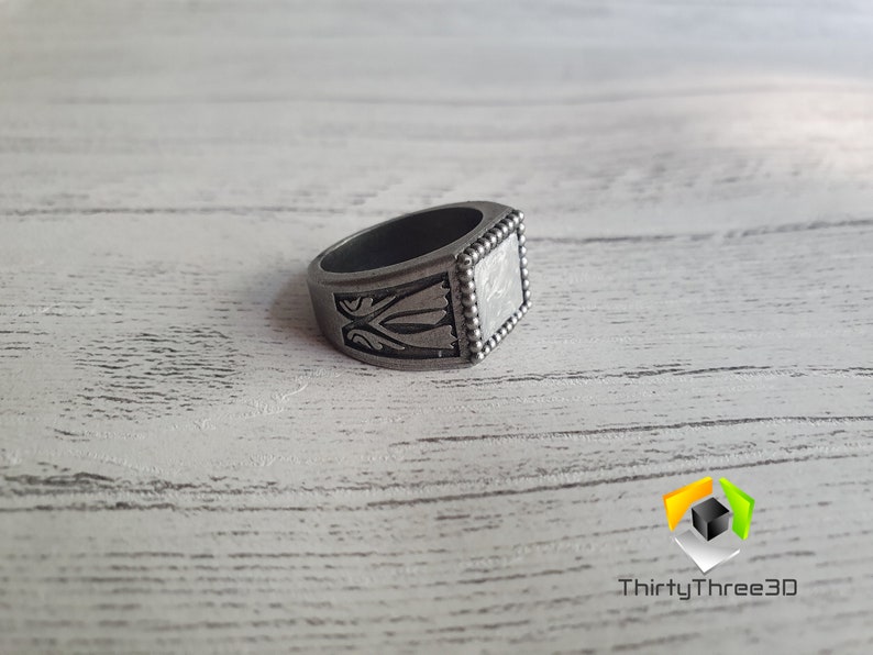 May include: A silver ring with a square face and intricate designs. The ring has a beaded edge and a textured surface.