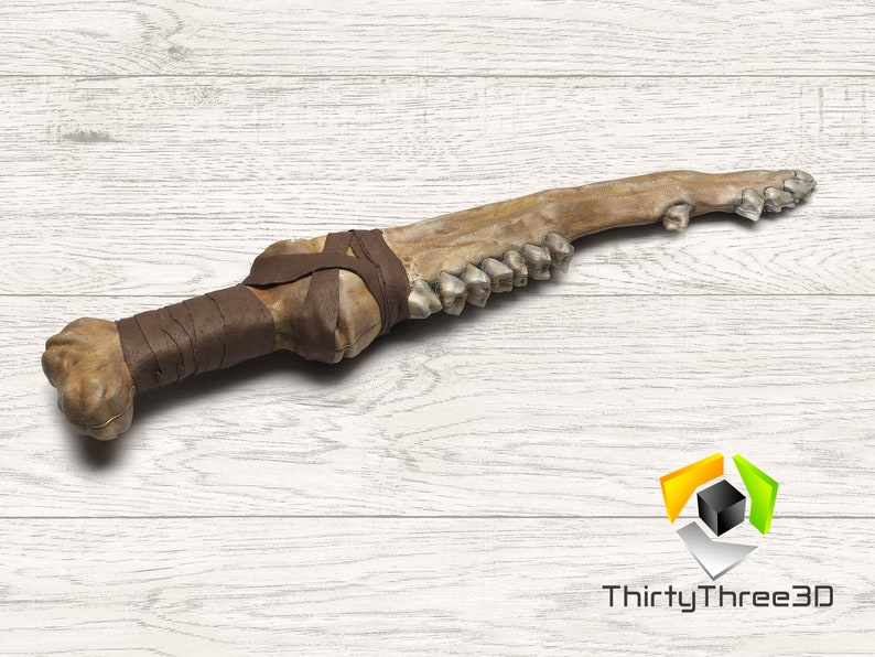 May include: A bone-like dagger with a wrapped handle and a row of teeth-like structures. The handle is wrapped with brown material. The dagger is set against a white wood-grain background. A unique, handcrafted item.