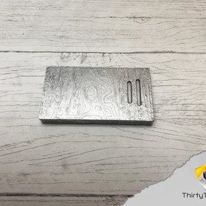 May include: A silver metal plate with a wood grain pattern and two rectangular cutouts. The plate is on a white wooden surface.