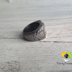May include: A silver ring with an intricate design on the top. The ring is lying on a white wooden surface.