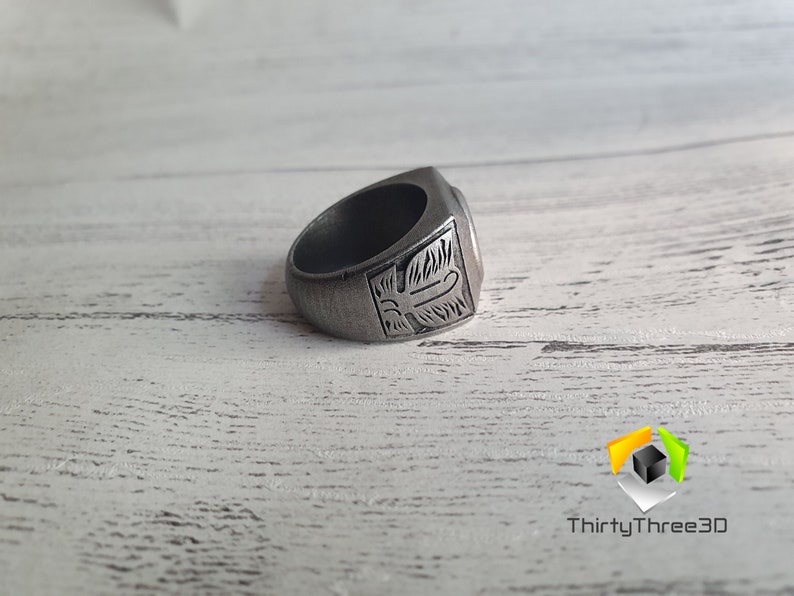 May include: A silver ring with an intricate design on the top. The ring is lying on a white wooden surface.