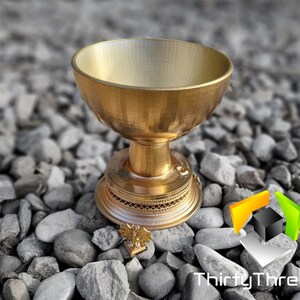 May include: A gold-colored metal cup with a decorative base, sitting on a bed of gray gravel stones. The cup has a smooth, rounded interior and a textured exterior. The base features intricate details and a small, gold-colored eagle design.