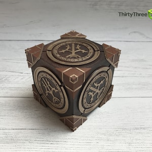 May include: A wooden cube with a brown finish and gold accents. Each side of the cube features a different design with a tree-like symbol. The cube is a decorative object.