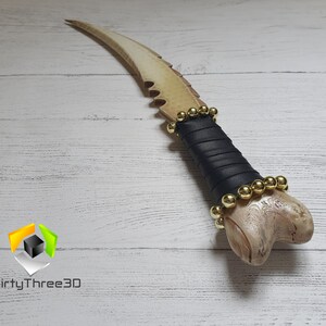 May include: A white bone-like dagger with a black leather wrap and gold beads. The dagger has a curved blade with a serrated edge.