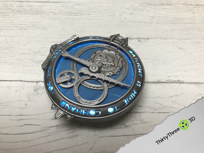 May include: A silver-colored circular pendant with a blue center and intricate gears. The inscription "TO RULE IS TO COMMAND" is engraved around the edge. The pendant is illuminated with a blue light.