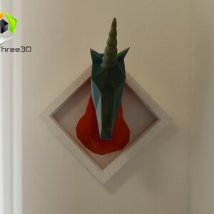 May include: A teal blue origami unicorn head mounted in a white square frame with a red background. The unicorn has a long, pointed horn.