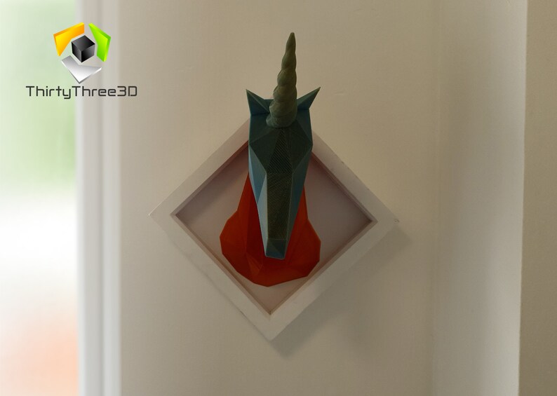May include: A teal blue origami unicorn head mounted in a white square frame with a red background. The unicorn has a long, pointed horn.