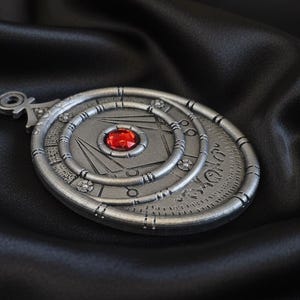 May include: A silver-colored pendant with a central red gemstone. The pendant features concentric circular designs and geometric patterns. The background is a dark, textured fabric, enhancing the pendant's details. The pendant has a loop at the top for a chain.
