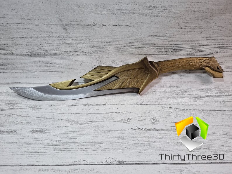May include: A gold and silver fantasy-style dagger with a detailed handle and a sharp blade. The dagger is lying on a white wooden surface.