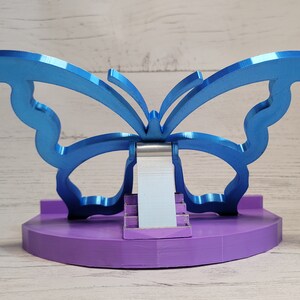 Peut inclure: Un support de t&eacute;l&eacute;phone en forme de papillon bleu avec une base violette. Le support est en m&eacute;tal et a un design moderne.