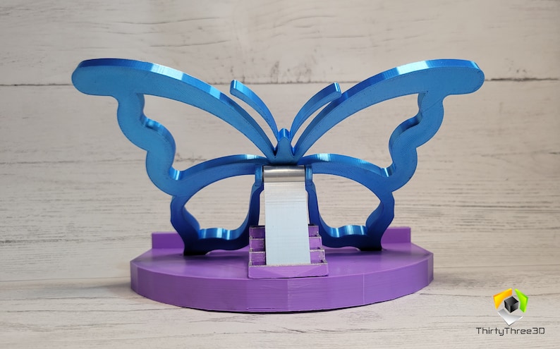 Peut inclure: Un support de t&eacute;l&eacute;phone en forme de papillon bleu avec une base violette. Le support est en m&eacute;tal et a un design moderne.