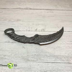 Puede incluir: Un cuchillo de metal plateado, curvo, con un patr&oacute;n decorativo en la hoja. El cuchillo tiene un anillo en la parte superior del mango.