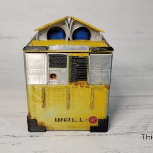 May include: A yellow and silver WALL-E robot toy with a rusty finish. The robot has a rectangular body with a small, square head and two blue eyes. The robot is standing on a white wooden surface.