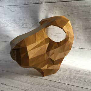 May include: A gold-colored, geometric mask with a single eye hole. The mask is made of a 3D printed material and has a textured surface.