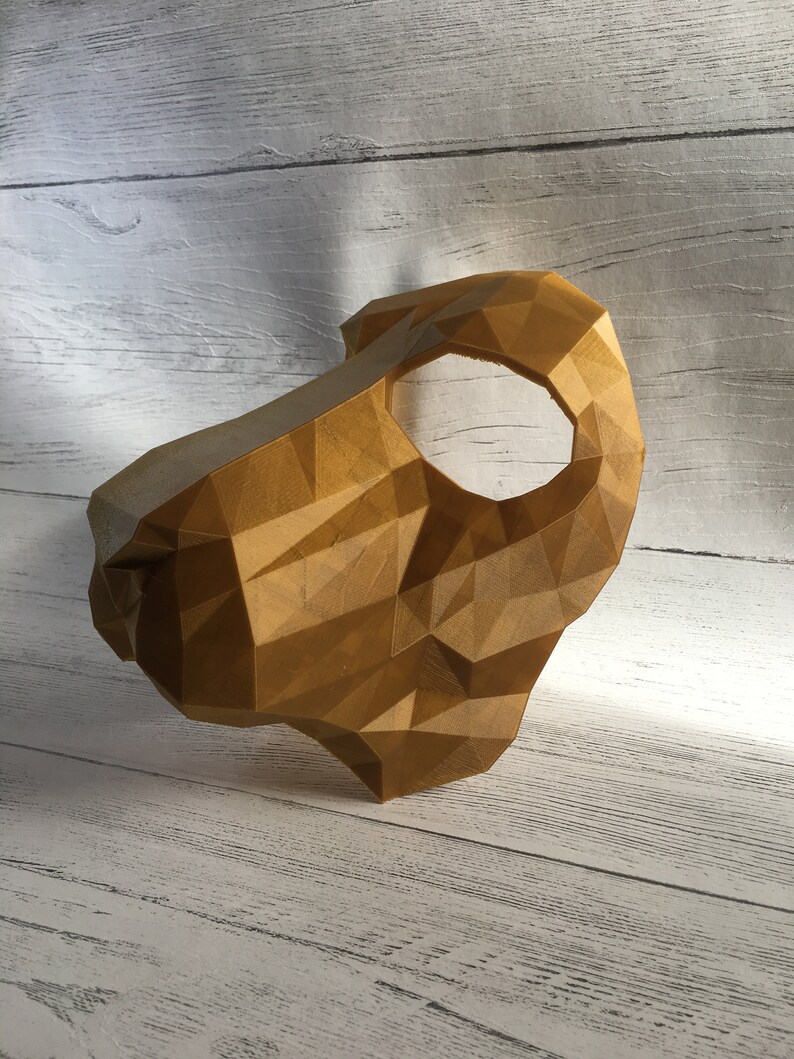 May include: A gold-colored, geometric mask with a single eye hole. The mask is made of a 3D printed material and has a textured surface.
