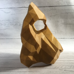 May include: A gold-colored, geometric mask with a triangular shape and a large eye hole. The mask is made of a 3D printed material and has a textured surface.