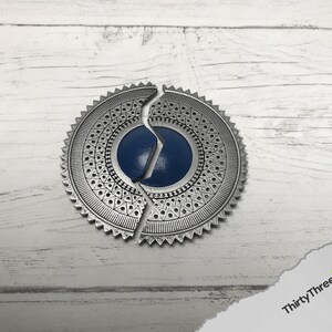 May include: A silver-colored metal medallion with a blue center, split into two pieces. The medallion has a decorative pattern around the edge.