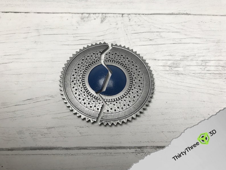 May include: A silver-colored metal medallion with a blue center, split into two pieces. The medallion has a decorative pattern around the edge.