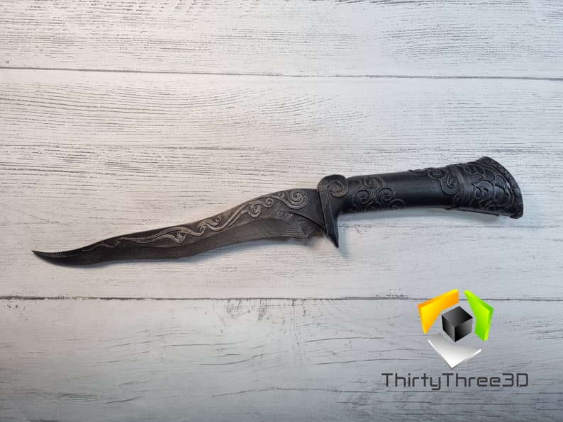May include: A black dagger with a curved blade and intricate floral designs on the handle. The dagger is made of metal and has a sharp point. The dagger is lying on a white wooden surface.