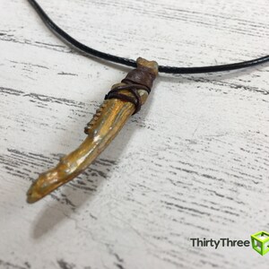 May include: A gold-colored necklace with a pendant shaped like a bone. The pendant is wrapped in brown leather and has a black cord.