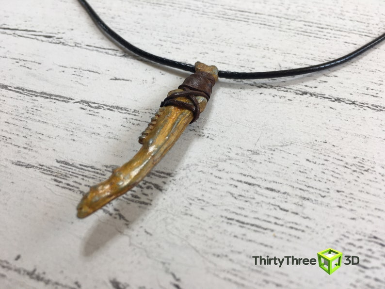 May include: A gold-colored necklace with a pendant shaped like a bone. The pendant is wrapped in brown leather and has a black cord.