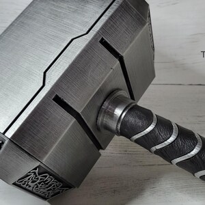May include: A silver-colored hammer with a black and silver handle. The hammer head has a detailed design and the handle has a textured grip.